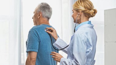Sarışın doktor hastanede steteskoplu olgun bir adamı muayene ediyor.