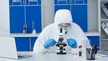 Tehlikeli madde giysisi içindeki virolog laboratuvarda dizüstü bilgisayarın yanında mikroskopla çalışıyor.