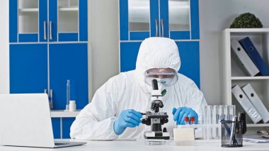 Tehlikeli madde giysisi içindeki bilim adamı test tüpleri ve dizüstü bilgisayarın yanında mikroskopla araştırma yapıyor.