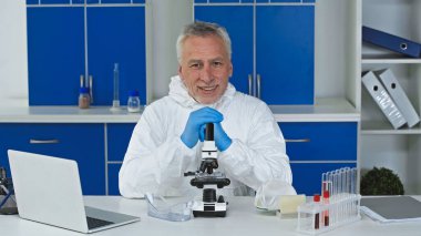 smiling scientist in hazmat suit looking at camera near microscope and laptop clipart