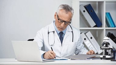Gözlüklü doktor iş yerinde laptopun yanına reçete yazıyor.