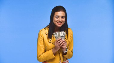 Cheerful young woman holding dollar banknotes isolated on blue clipart