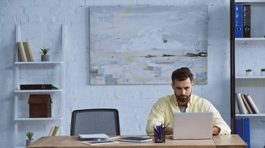 Gündelik kıyafetlerle genç yönetici ofiste dizüstü bilgisayarda çalışıyor.