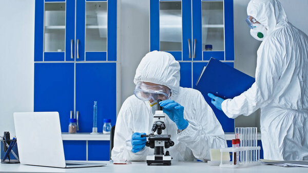 virologist working with microscope while colleague holding clipboard in laboratory