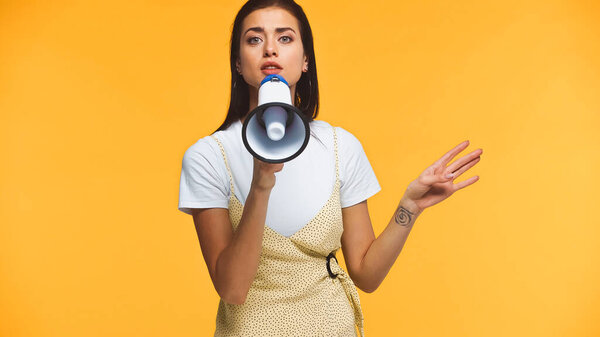 young woman talking at loudspeaker and pointing with hand isolated on yellow