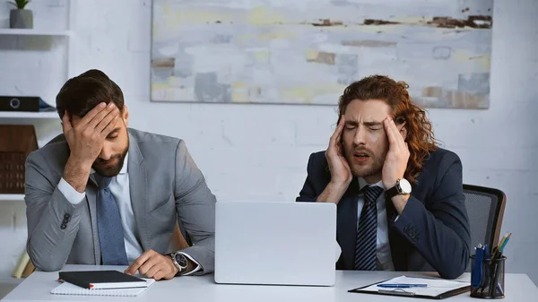 Vyčerpaní Podnikatelé Dotýkají Hlavy Zatímco Sedí Blízkosti Notebooku Kanceláři — Stock fotografie