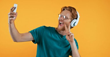 Kablosuz kulaklıklı şaşkın kızıl saçlı adam sarı renkte barış işareti gösterirken selfie çekiyor. 