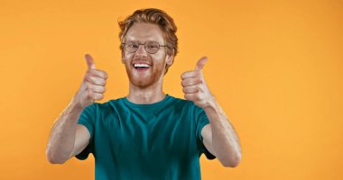 cheerful redhead man in glasses showing thumbs up isolated on yellow  clipart