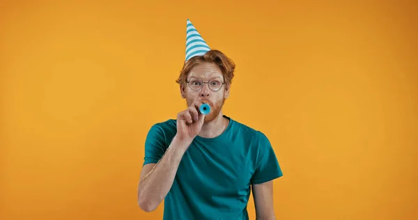 redhead man in party cap blowing in party horn isolated on yellow