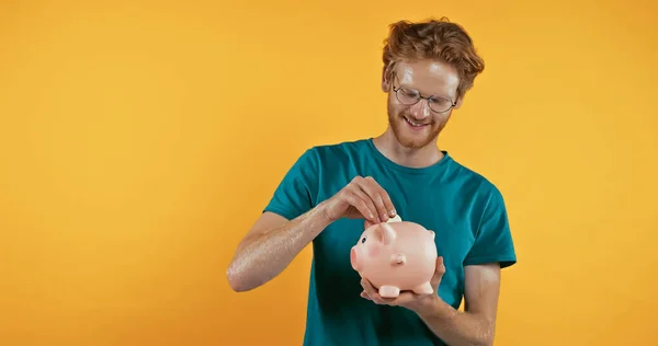 Happy Redhead Man Putting Coin Piggy Bank Isolated Yellow — Stock Photo, Image