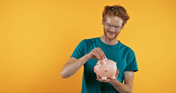 happy redhead man putting coin in piggy bank isolated on yellow 