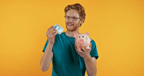 positive man holding piggy bank and dollars isolated on yellow