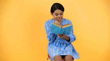 amazed schoolgirl in blue dress reading book while sitting isolated on yellow clipart