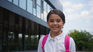 smiling schoolgirl with backpack standing near building outside clipart