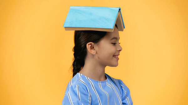 happy child with book on head smiling isolated on yellow