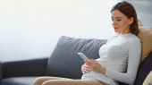 mladá těhotná žena sedí na gauči a zprávy na smartphone