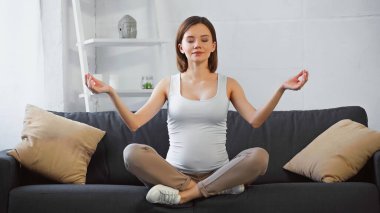 Genç hamile kadın, evdeki kanepede gözleri kapalı meditasyon yapıyor.