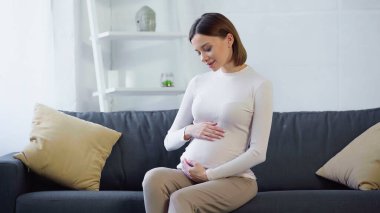 Gülümseyen, hamile bir kadın evdeki koltukta otururken karnına dokunuyor.