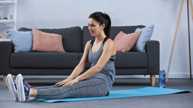 Spor kıyafetli genç bir kadın evde spor minderinde oturuyor. 