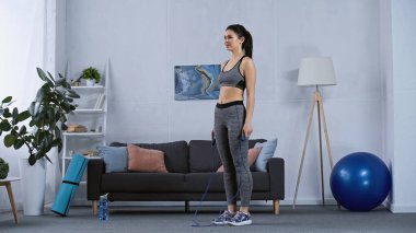 Spor giyimli genç bir kadın atlama halatıyla evde çalışıyor. 