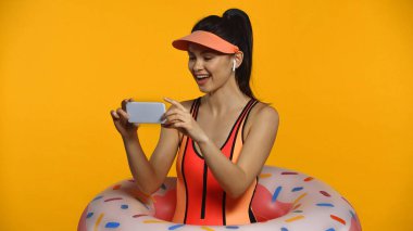 Kablosuz kulaklıklı, mayolu ve şişme yüzüklü neşeli genç bir kadın sarıda izole edilmiş akıllı telefon kullanıyor.