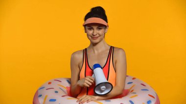 Cheerful young woman in swimsuit and inflatable ring holding megaphone isolated on yellow clipart