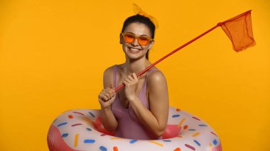 Cheerful young woman in swimsuit and inflatable ring holding scoop-netisolated on yellow clipart
