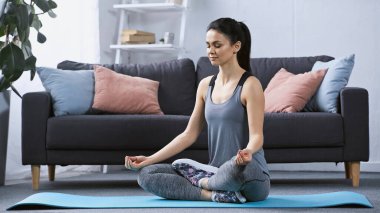 Spor kıyafetli genç bir kadın evde fitness minderi üzerinde meditasyon yapıyor. 