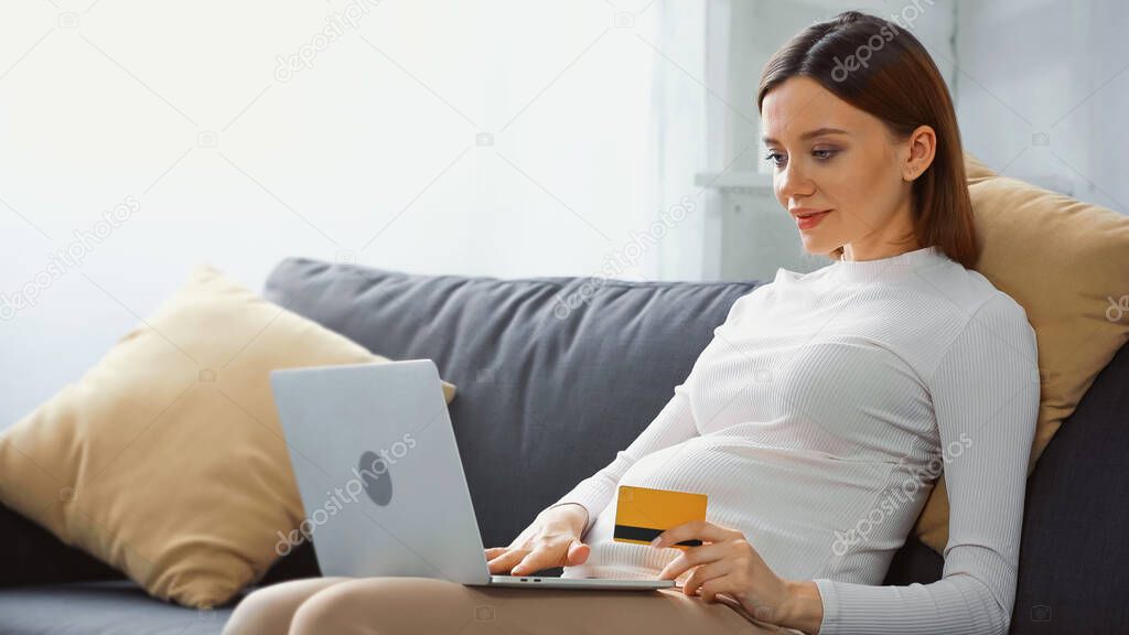 pretty, pregnant woman holding credit card while using laptop on couch