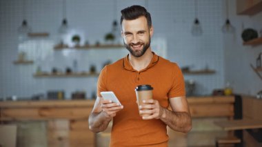 Gülümseyen adam akıllı telefon kullanıyor ve kafeye gitmek için kahve tutuyor.