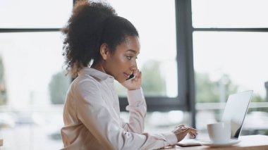 Kafede dizüstü bilgisayar kullanırken akıllı telefondan konuşan Afro-Amerikan kadının yan görüntüsü 