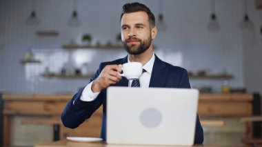 Kahvede dizüstü bilgisayarın yanında elinde kahve tutan takım elbiseli memnun bir adam.