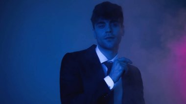 Takım elbiseli adam dumanlı mavi kameraya bakıyor.