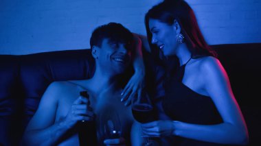 Mutlu ve gömleksiz bir adam elinde şarap şişesiyle seksi bir kız arkadaşın yanında mavinin üstünde