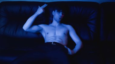 Siyah kanepede yatarken pantolonlu, poz veren ve el kol hareketi yapan üstsüz bir adamın üzerinde mavi ışık. 