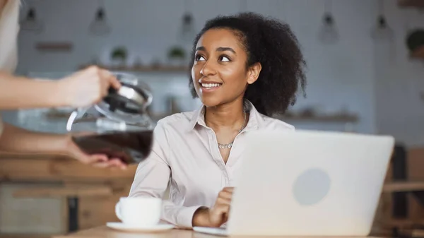 happy african american businesswoman looking at waitress with coffee pot in cafe