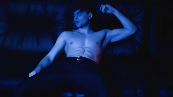 shirtless man in pants relaxing on black sofa on blue