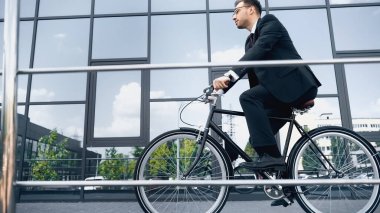 full length of businessman in suit and glasses riding bike near building with glass facade clipart