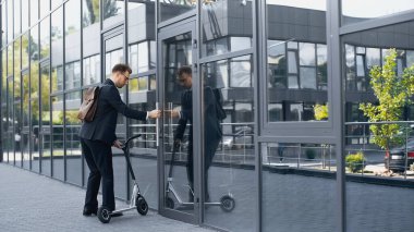 Takım elbiseli ve sırt çantalı bir işadamı elektrikli scooterla binaya giriyor. 