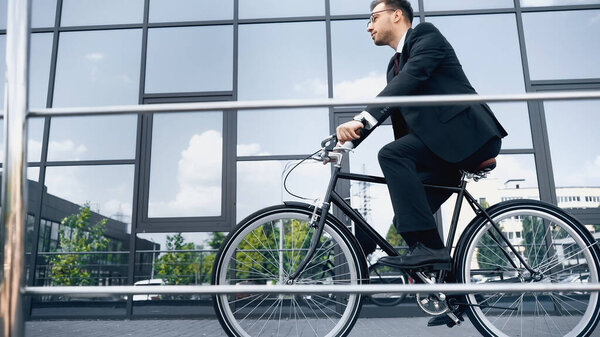 full length of businessman in suit and glasses riding bike near building with glass facade