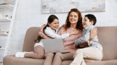 Usmívající se matka objímající děti v blízkosti notebooku na gauči 