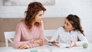 Kalemi olan çocuk evde defterin yanında annesiyle konuşuyor. 