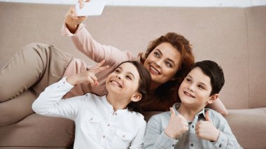 Neşeli çocuklar evde gülümseyen anneleriyle selfie çekerken el kol hareketi yapıyorlar. 