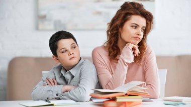 Displeased woman and son sitting near notebook and books at home  clipart