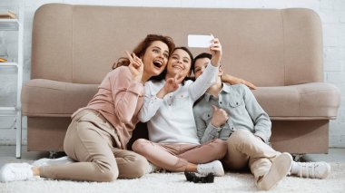 KYIV, UKRAINE - 15 Nisan 2019: Evinde yerde selfie ve av alanı alan çocukların yanında barış işareti gösteren neşeli kadın 