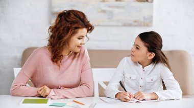 Yanında oturan gülümseyen kadın deftere ve yeşil ekranlı dijital tablete yazı yazıyor. 