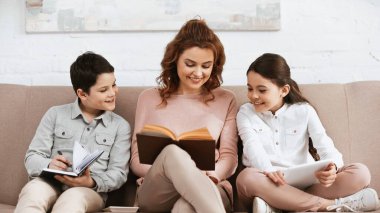 Elinde not defteri ve dijital tabletle çocukların yanında kitap okuyan gülümseyen kadın. 