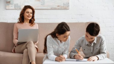 Gülümseyen tele-işçi bulanık ön planda ödev yapan çocuklara bakıyor. 