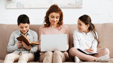 Kanepede laptopla annelerinin yanında ödev yapan gülümseyen çocuklar. 