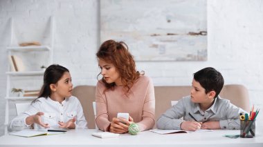 Annemin yanındaki defteri işaret eden kız akıllı telefonlu ve erkek kardeşi oturma odasında. 